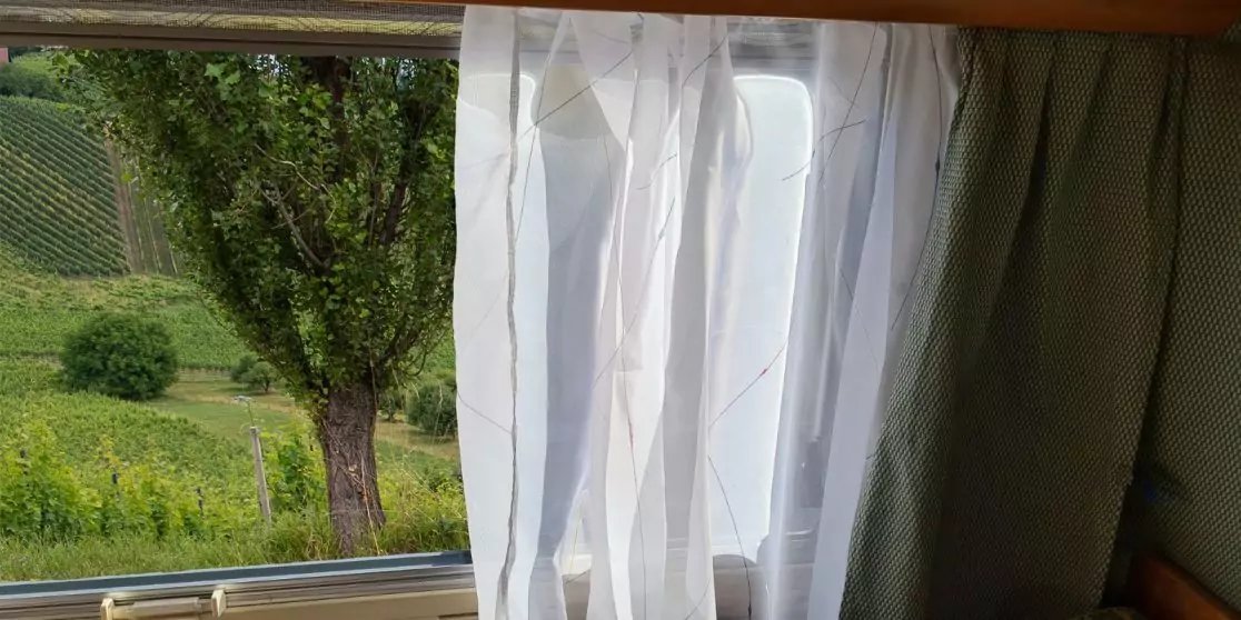 Fenster mit halbtransparentem wei&szlig;em Vorhang, rechts gr&uuml;ner Vorhang; drau&szlig;en Grasfl&auml;che, ein Baum und ein H&uuml;gel sichtbar