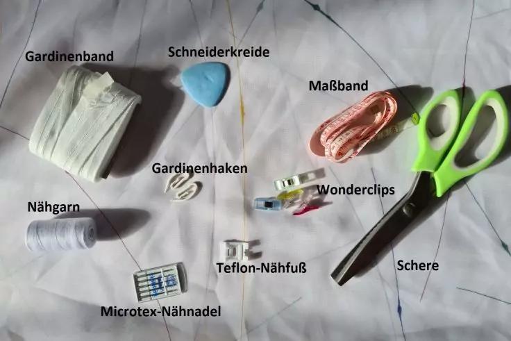 Verschiedene N&auml;hutensilien auf wei&szlig;em Stoff: Gardinenband, Schneiderkreide, Ma&szlig;band, gr&uuml;ne Schere, N&auml;hgarn, Gardinenhaken, Wonderclips, Teflon‑N&auml;hfu&szlig; und Microtex‑N&auml;hnadel