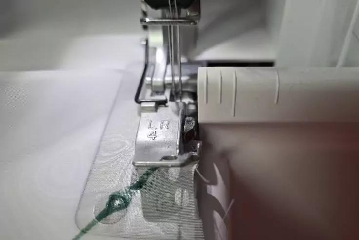 Nahaufnahme einer N&auml;hmaschine mit Metall-N&auml;hfu&szlig;, Nadel, wei&szlig;em Stoff mit gr&uuml;ner Naht und wei&szlig;em Kunststoffteil rechts