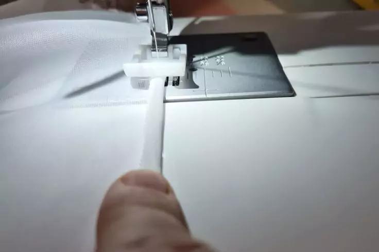 N&auml;hmaschine mit N&auml;hfu&szlig; und Nadel &uuml;ber wei&szlig;em Stoff, Finger h&auml;lt Stoffkante