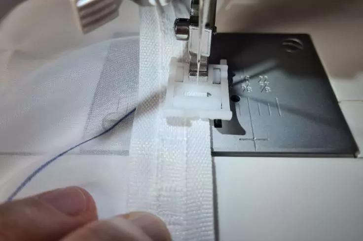 Nahaufnahme einer N&auml;hmaschine: Nadel und N&auml;hfu&szlig;, wei&szlig;e Stoffkante mit Naht und zwei Finger, die das Material f&uuml;hren