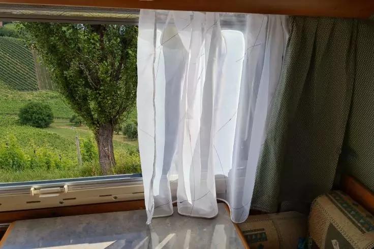 Wei&szlig;e, transparente Gardine h&auml;ngt vor einem ge&ouml;ffneten Fenster; gr&uuml;ner Vorhang zur Seite; Blick auf Baum und Wiese; Holzrahmen; marmorierte Tischplatte im Vordergrund; gepolsterter Sitz rechts