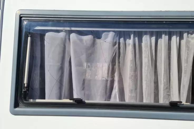 Rechteckiges Fenster mit hellgrauem Rahmen, geschlossenen wei&szlig;en Vorh&auml;ngen mit Falten und zwei schwarzen Verschl&uuml;ssen unten