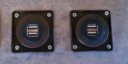 Zwei schwarze quadratische Einbaurahmen nebeneinander, jeweils mit zwei &uuml;bereinander liegenden USB-A-Anschl&uuml;ssen, vier Schrauben an den Ecken, auf grauer Betonoberfl&auml;che