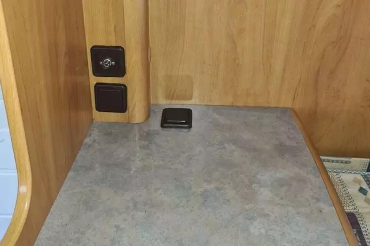 Graue Tischplatte vor Holzvert&auml;felung; links an einem Holzpfosten zwei schwarze Steckdosen; auf der Platte ein kleines schwarzes rechteckiges Ger&auml;t; rechts der Rand eines gemusterten Tuchs