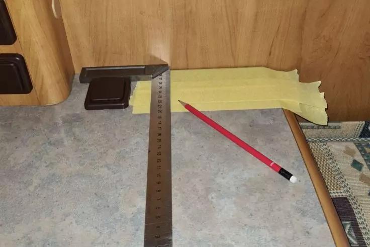 Arbeitsfl&auml;che vor Holzverkleidung mit Metalllineal, rotem Bleistift, schwarzem rechteckigem Ger&auml;t, gelbem Klebeband an der R&uuml;ckkante und gemustertem Stoff rechts