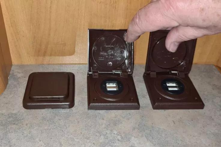 Drei braune quadratische Plastikschachteln auf einer Arbeitsfl&auml;che; zwei ge&ouml;ffnet, jeweils mit rundem Fach und kleinem schwarzen Ger&auml;t mit rechteckigem Display; linke Schachtel geschlossen; Hand zeigt auf die rechte ge&ouml;ffnete Schachtel