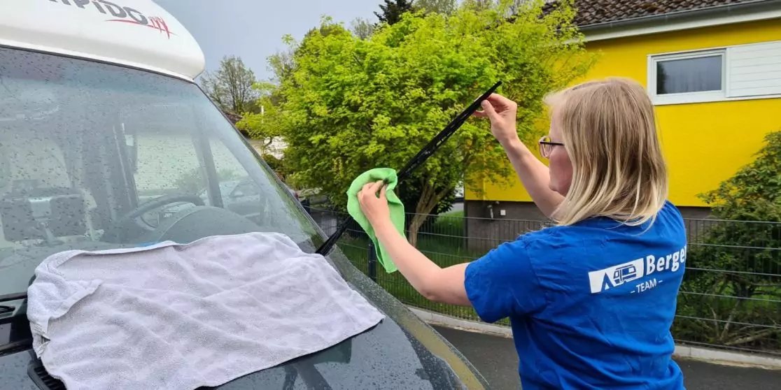 Person mit blondem Haar reinigt die Windschutzscheibe eines wei&szlig;en Vans mit Abzieher und gr&uuml;nem Tuch; wei&szlig;e Decke auf der Motorhaube, gelbes Haus und gr&uuml;ner Busch im Hintergrund