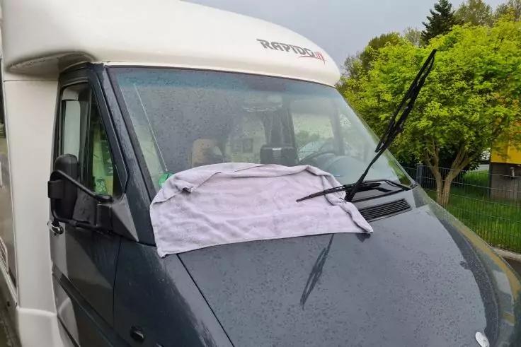 Vorderansicht eines wei&szlig;en Wohnmobils, ein helles Handtuch liegt auf der Windschutzscheibe, ein Scheibenwischer ist angehoben, Regentropfen auf der Motorhaube