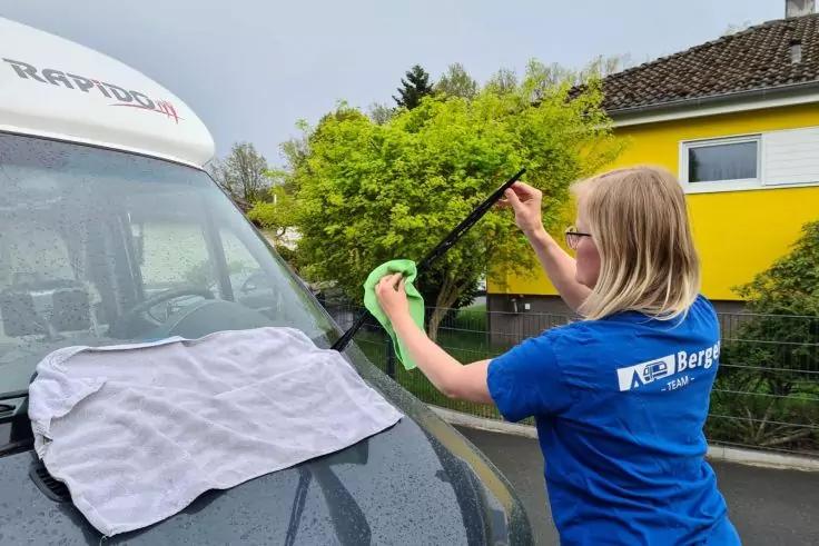 Person mit blondem Haar und blauem T‑Shirt reinigt mit einem gr&uuml;nen Tuch einen hochgeklappten Scheibenwischer an der Front eines dunkelgrauen Fahrzeugs; ein graues Tuch liegt auf der Motorhaube, im Hintergrund Haus und Str&auml;ucher