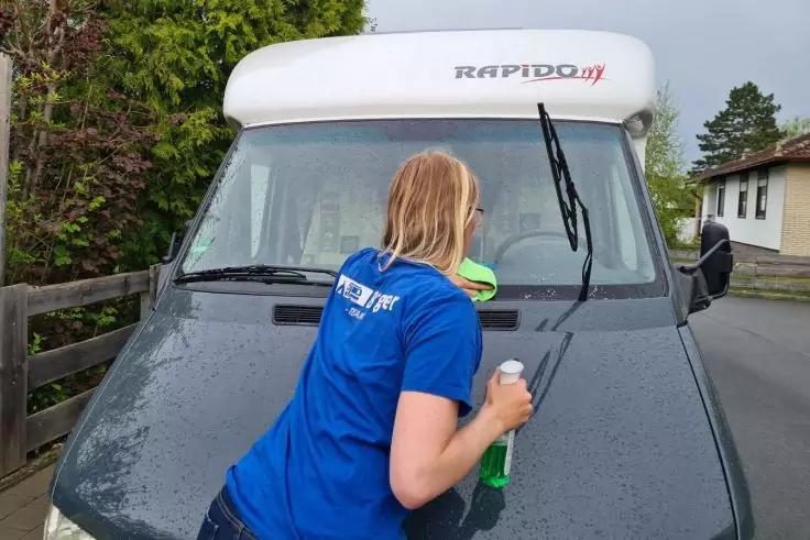 Person mit schulterlangen blonden Haaren und blauem T‑Shirt steht vor der Windschutzscheibe eines wei&szlig;en Wohnmobils mit Schriftzug "RAPIDO", h&auml;lt eine gr&uuml;ne Spr&uuml;hflasche und ein Tuch an der Scheibe