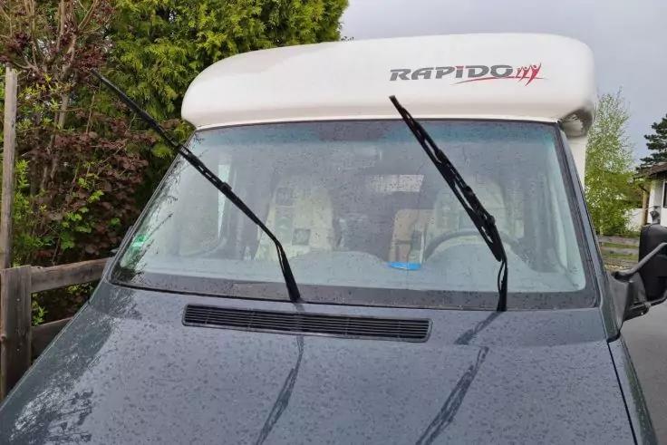 Frontansicht eines grauen Wohnmobils mit dem Schriftzug RAPIDO auf dem Aufbau, hochgestellte Windschutzscheibenwischer, Regentropfen auf der Scheibe, links Holzzaun und B&auml;ume