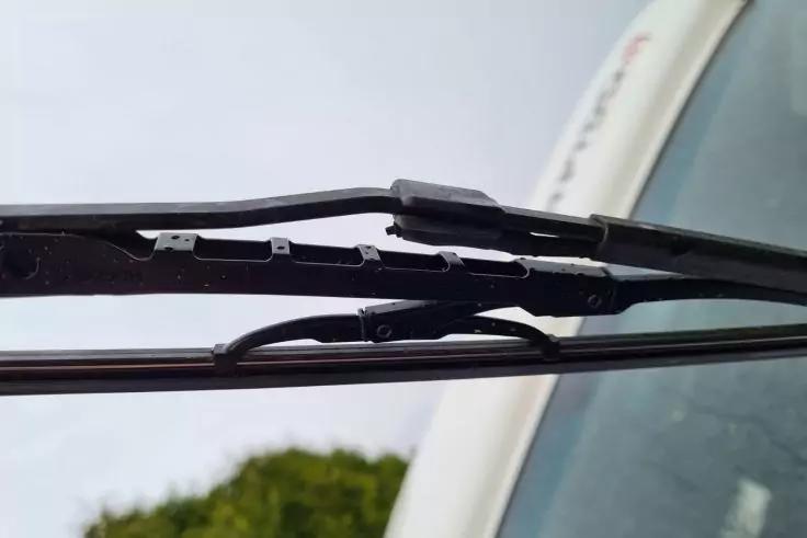 Nahaufnahme von zwei Scheibenwischern auf einer Windschutzscheibe, Himmel und gr&uuml;nes Geb&uuml;sch im Hintergrund