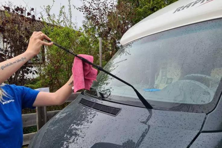Teilweise sichtbarer Arm in blauem T-Shirt reinigt mit einem pinken Tuch den hochgeklappten Scheibenwischer eines grauen Vans; regennasse Windschutzscheibe und Haube; Pflanzen im Hintergrund