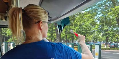 Person von hinten mit blondem Pferdeschwanz, tr&auml;gt blaues T-Shirt, streckt den Arm aus dem Fahrzeugsfenster und h&auml;lt ein rotes Objekt; B&auml;ume und Parkschranken im Hintergrund