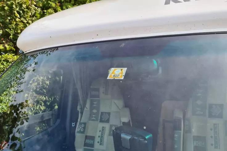 Frontscheibe eines wei&szlig;en Fahrzeugs mit kleinem gelbem quadratischen Aufkleber oben in der Mitte, zwei gepolsterte Sitze mit gemusterten Sitzbez&uuml;gen, ein schwarzes kastenf&ouml;rmiges Ger&auml;t auf dem Armaturenbrett und Spiegelung von gr&uuml;nem Laub links