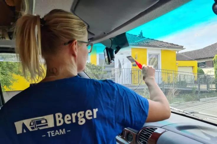Person mit blondem Pferdeschwanz in blauem Shirt mit Schriftzug Berger, sitzt in einem Fahrzeug und h&auml;lt einen roten Abzieher an die Windschutzscheibe; Arm, Armaturenbrett und ein gelbes Haus mit Zaun sind sichtbar