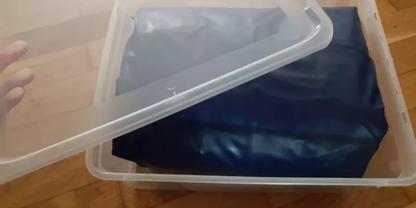 Transparente Kunststoffbox mit halb ge&ouml;ffnetem Deckel, darin ein zusammengelegtes dunkelblaues Stoffst&uuml;ck; Holzfu&szlig;boden sichtbar, linke Hand h&auml;lt den Deckel