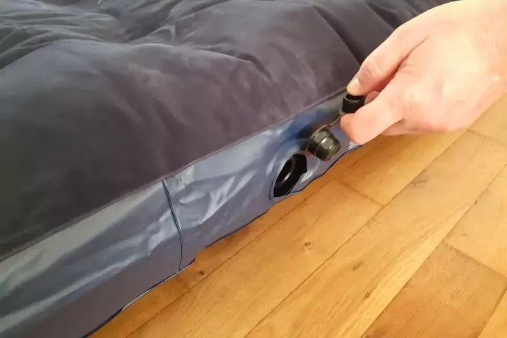 Hand h&auml;lt schwarzes Ventil an einer grauen aufblasbaren Luftmatratze auf einem Holzboden