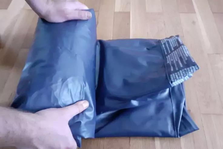 Zwei H&auml;nde und ein Fu&szlig; rollen eine schwarze Plastikt&uuml;te auf einem hellen Holzfu&szlig;boden; daneben gefaltete schwarze Plastikt&uuml;ten mit wei&szlig;er Aufschrift