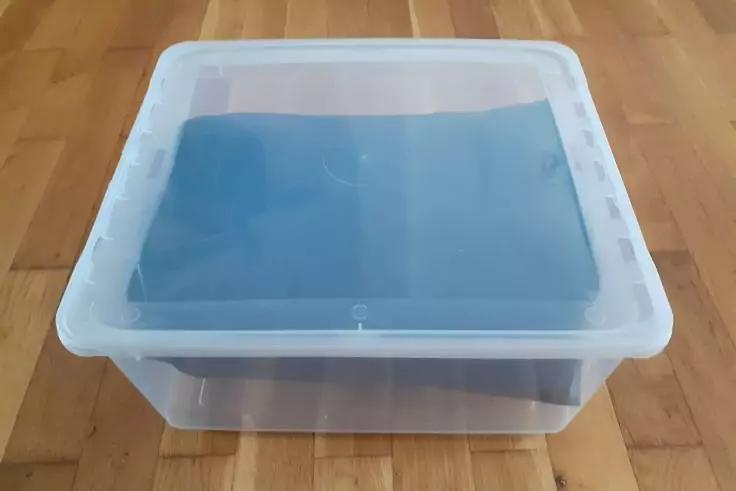Transparente rechteckige Plastikbox mit wei&szlig;em Deckel, darin gefaltetes blaues Textil, auf Holzfu&szlig;boden