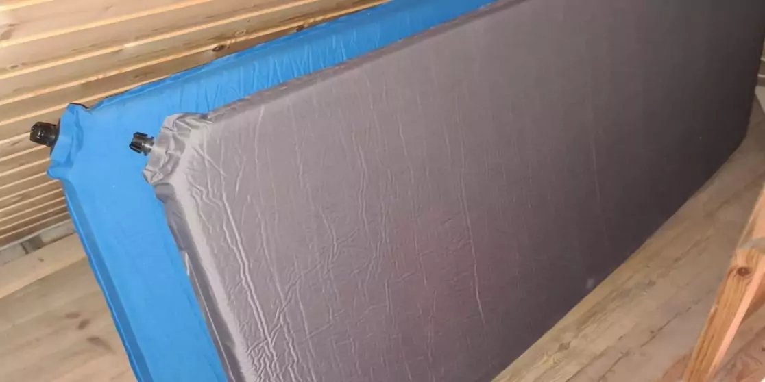 Zwei aufrecht an eine Holzlattenwand gelehnte Matratzen auf Holzfu&szlig;boden; vorn grau bezogen, hinten blau mit sichtbarem Ventil