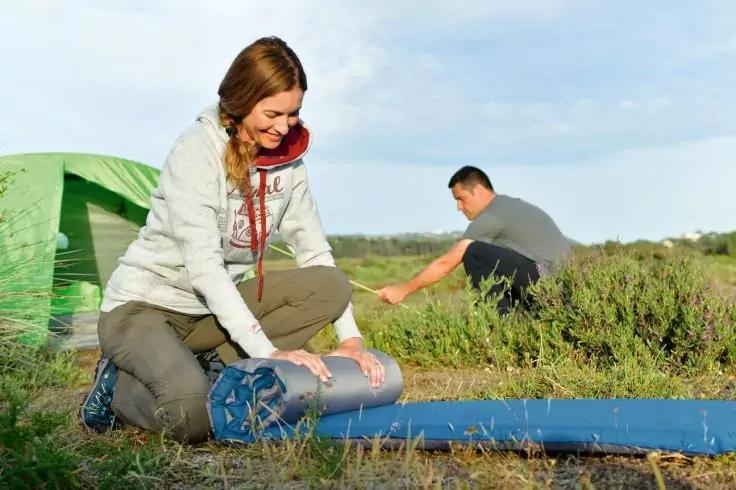 Frau kniet und rollt eine Isomatte neben einem gr&uuml;nen Zelt; ein Mann im Hintergrund beugt sich nach vorn; Gras und wolkiger Himmel