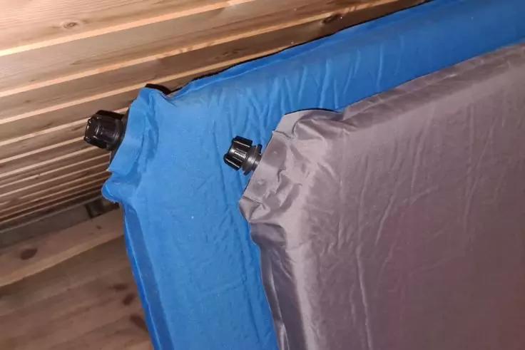 Zwei aufblasbare Luftmatratzen, eine blau, eine grau, mit schwarzen Ventilverschl&uuml;ssen, lehnen nebeneinander an einer Holzlattenwand