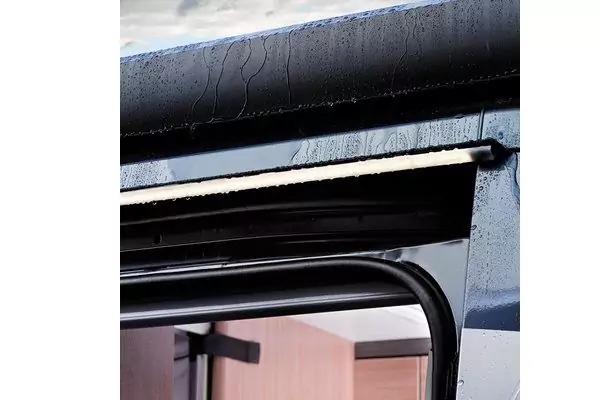 Nahaufnahme einer geöffneten Türöffnung mit schwarzem Gummidichtungsrahmen und Metallprofil, Wassertropfen auf der Oberkante, hellbraune Holzverkleidung im Innenraum, grauer Himmel oben