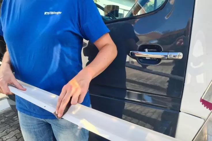 Oberk&ouml;rper einer Person in blauem T‑Shirt, die eine lange wei&szlig;e Leiste vor einer dunklen Fahrzeugt&uuml;r mit sichtbarem T&uuml;rgriff h&auml;lt