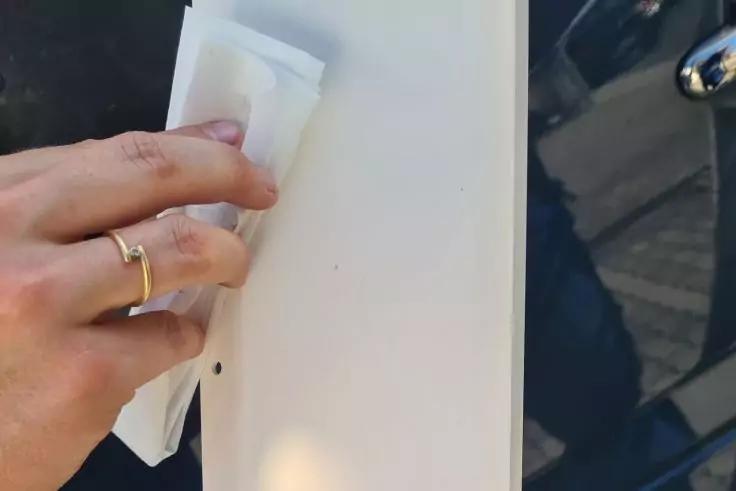 Hand mit Goldring h&auml;lt ein wei&szlig;es Tuch an eine wei&szlig;e vertikale Platte; rechts eine dunkle gl&auml;nzende Fl&auml;che