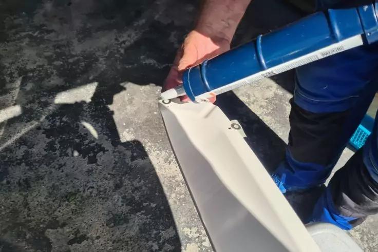 H&auml;nde halten eine Kartuschenpistole mit blauer Kartusche und tragen wei&szlig;e Dichtmasse auf ein wei&szlig;es Kunststoffprofil auf; grauer Boden und blaue Arbeitskleidung sichtbar