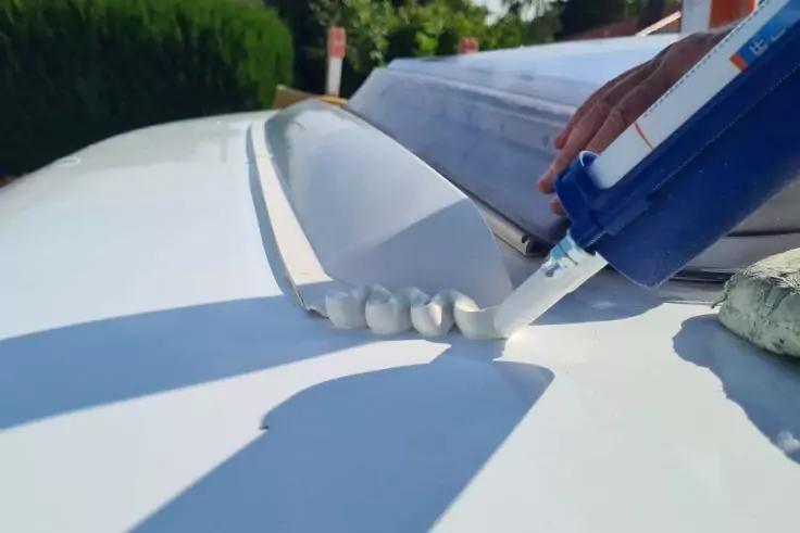 Hand mit blauer Kartuschenpistole, die wei&szlig;e Dichtmasse in Perlen auf eine wei&szlig;e Fl&auml;che auftr&auml;gt; im Hintergrund unscharfe Vegetation und ein Schild