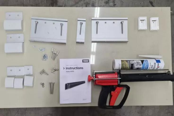 Auf einer hellen Fl&auml;che liegen wei&szlig;e Metallplatten und Winkel mit L&ouml;chern, mehrere Schrauben und D&uuml;bel, eine Bedienungsanleitung sowie eine schwarze Kartuschenpresse mit rotem Griff und einer Tube