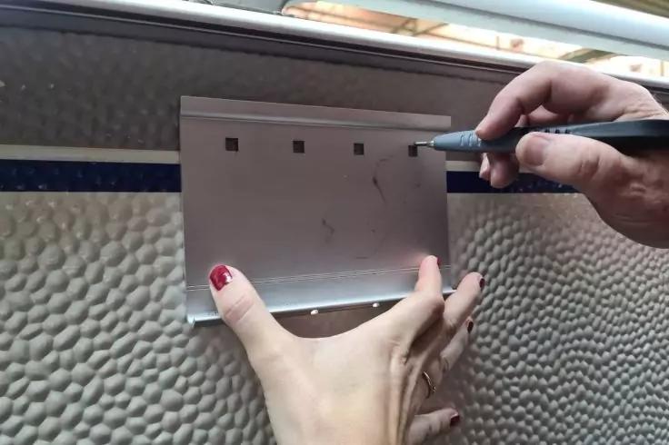 Zwei H&auml;nde halten ein rechteckiges wei&szlig;es Kunststoffteil an einer strukturierten Wand; die rechte Hand h&auml;lt einen schwarzen Marker, auf dem Kunststoff sind Markierungen; die linke Hand zeigt roten Nagellack und einen Ring