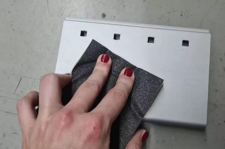 Linke Hand mit roten Fingern&auml;geln h&auml;lt schwarzen Schleifschwamm an wei&szlig;em rechteckigem Gegenstand mit vier quadratischen &Ouml;ffnungen auf grauem Untergrund