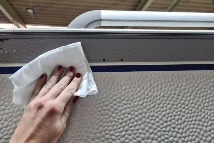 Hand mit roten Fingern&auml;geln h&auml;lt ein wei&szlig;es Tuch gegen eine strukturierte hellgraue Oberfl&auml;che unter einer Metallleiste