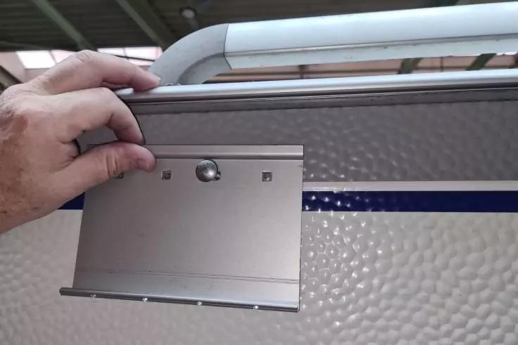 Hand h&auml;lt eine rechteckige Metallplatte mit sichtbaren Schrauben vor einer wei&szlig;en, genoppten Fl&auml;che mit blauem Streifen und einem gebogenen wei&szlig;en Metallb&uuml;gel oben