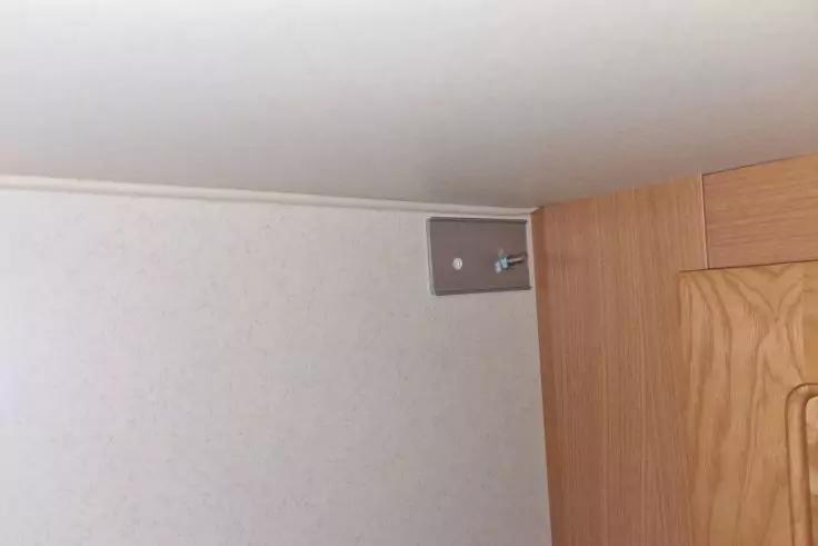 Ecke einer Zimmerdecke und -wand; rechts ein Holzm&ouml;bel; an der Wand nahe der Decke eine kleine rechteckige Metallplatte mit zwei Schrauben und einem wei&szlig;en Kunststoffteil