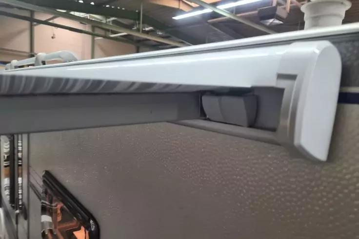 Wei&szlig;e, zylindrische aufgerollte Bahn in einer wei&szlig;en Halterung, montiert an einer hellgrauen, genoppten Au&szlig;enwand; links unten ein schwarzer Fensterrahmen mit orangefarbener Innenfl&auml;che; oben sichtbare Deckenrohre und Leuchten