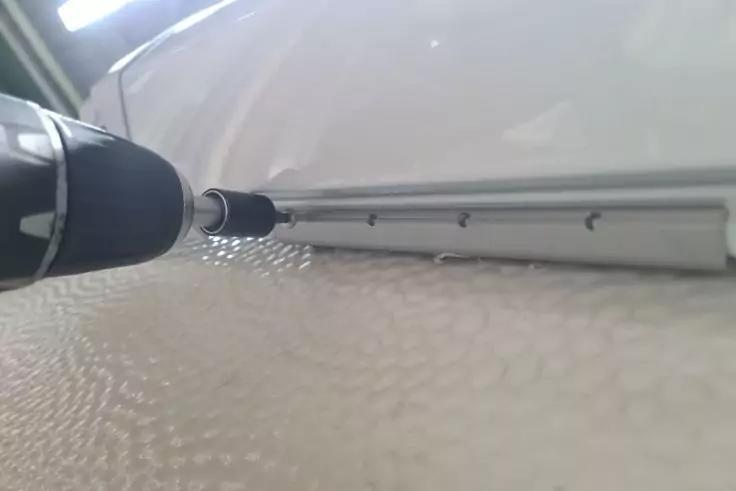 Schwarzer Akkuschrauber-Bit treibt eine Schraube in eine silberne Metallleiste mit mehreren Schrauben; dar&uuml;ber eine wei&szlig;e glatte Fl&auml;che, darunter eine strukturierte wei&szlig;e Oberfl&auml;che