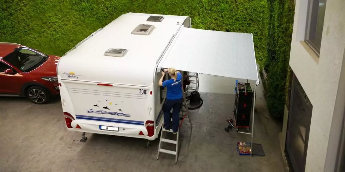 Wei&szlig;es Wohnmobil mit ausgefahrener Markise; eine Person auf einer Leiter neben dem Fahrzeug; rotes Auto, hohe gr&uuml;ne Hecke, Geb&auml;udefassade mit Fenster, Gegenst&auml;nde auf dem Boden