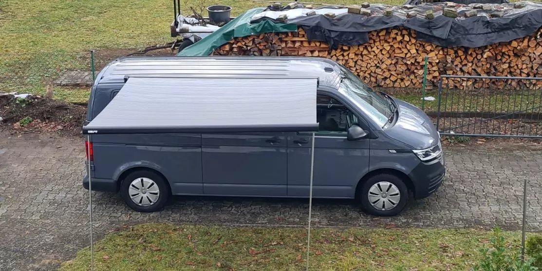 Grauer Transporter seitlich auf gepflasterter Fahrspur geparkt, im Hintergrund gestapelte Holzscheite unter schwarzer Plane, Grasfl&auml;chen daneben; keine Personen sichtbar