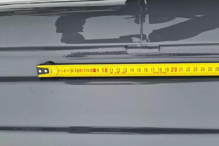 Gelbes Maßband, ausgezogen bis etwa 24 cm, liegt quer auf dunkler, geriffelter Metallfläche; darüber ein Scharnier und die Reflexion einer Person und einer Hand