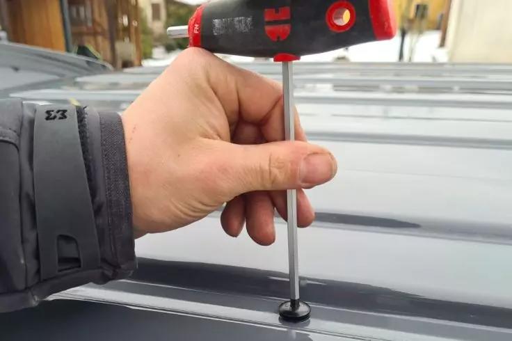 Hand in grauer Jacke hält roten Schraubendreher und dreht eine Schraube in eine graue Metallplatte