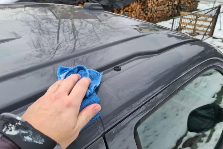 Hand mit blauem Tuch auf dem Dach eines dunkelgrauen Autos; Dach zeigt Baumreflexionen; Seitenfenster und Außenspiegel sichtbar; schneebedeckter Boden und Holzstapel im Hintergrund