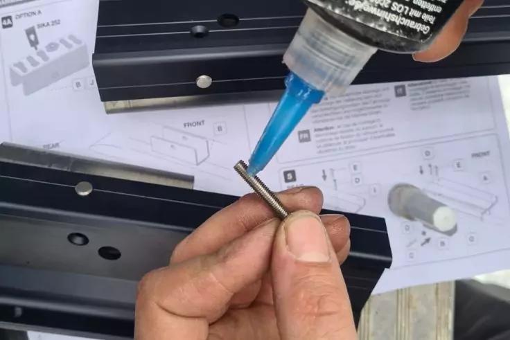 Zwei Hände halten einen kurzen Metallstift, eine Flasche mit blauer Düse gibt Klebstoff auf das Ende; im Hintergrund schwarze Metallprofile und bedruckte Papiere mit Diagrammen