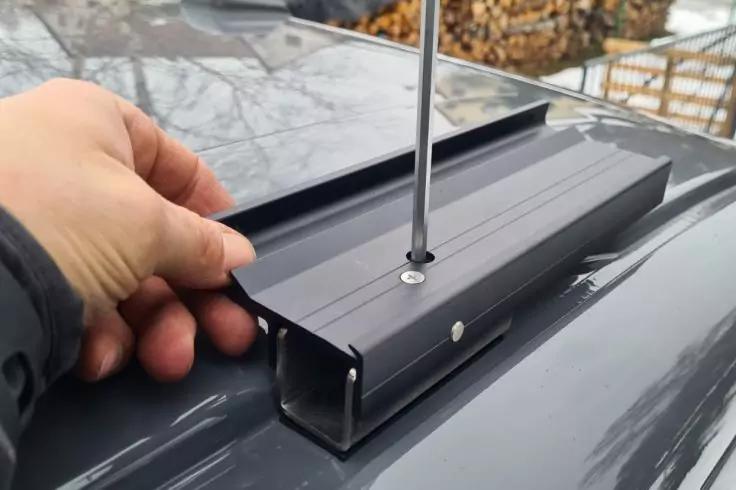 Hand hält schwarzes Metallprofil mit senkrechtem Schraubenschaft auf glänzendem dunkelgrauem Autodach; im Hintergrund gestapeltes Brennholz und Schnee