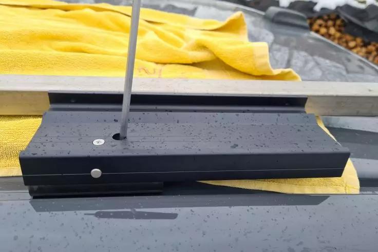 Schwarzes rechteckiges Bauteil auf nassem Autodach mit Wassertropfen, teilweise bedeckt von einem gelben Tuch; eine dünne Metallstange steckt in einer Schraube