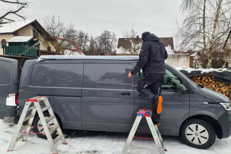 Grauer Transporter steht im Schnee vor Wohnhäusern; Person in schwarzer Kapuzenjacke mit einem Fuß auf dem Fahrzeug und einem Fuß auf einer Leiter; zwei Aluleitern, Holzhaufen und kahle Bäume im Hintergrund
